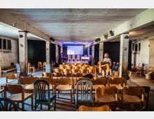 Ein einfacher Veranstaltungsraum mit unbehandelten Wänden, mehreren Holzsäulen und einer kleinen Bühne mit Mikrophonen und Stühlen. Im Vordergrund stehen zahlreiche Holzstühle, einige Personen richten den Raum ein.