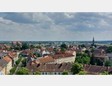 Stadtansicht mit roten Ziegeldächern und einer Kirche mit spitzem Turm im Hintergrund, umgeben von grünen Bäumen. Im Hintergrund sind flache Felder, eine Brücke und ein bewölkter Himmel zu sehen.