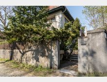 Wohnhaus mit Garten in einer ruhigen Straße, umgeben von Bäumen. Sichtbar ist ein Eingangstor aus Stein mit Treppen, die zu einem zweistöckigen Gebäude mit hellem Putz und roten Ziegeldach führen.