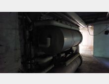 Dunkler Kellerraum mit einer großen metallischen Zylinderanlage, verbunden durch Rohre und Leitungen. Im Hintergrund ist ein heller, leerer Raum mit offener Tür zu sehen.