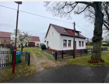 Ein freistehendes weißes Einfamilienhaus mit rotem Satteldach und roten Fensterrahmen steht auf einem eingezäunten Grundstück. Im Vordergrund sind ein blühender Baum, eine Einfahrt mit unbefestigtem Weg und ein Nebengebäude mit ziegelrotem Dach sichtbar.
