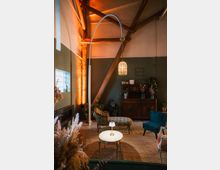 Gemütliches Wohnzimmer mit einer rustikalen Holzbalkendecke, grünen Wänden und warmen Lichtakzenten. Im Zentrum stehen eine antike Kommode, ein kleines Tischchen mit Lampe, gepolsterte Sessel und ein Dekorationsobjekt in Form einer Gorilla-Figur.