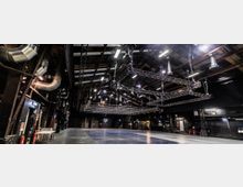 Innenansicht einer großen, industriell gestalteten Veranstaltungshalle mit freiliegenden Rohren und Metallträgern an der Decke. Im Raum befinden sich hängende Beleuchtungsgerüste, dunkle Wände und mehrere technische Elemente an den Seiten.