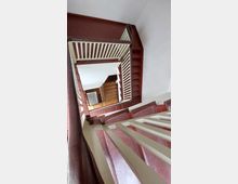 Treppe in einem mehrstöckigen Gebäude mit rot gestrichenen Holzstufen, weißen Geländern und einem rechteckigen Treppenauge. Unten ist ein Holzboden sichtbar, der den unteren Treppenbereich markiert.