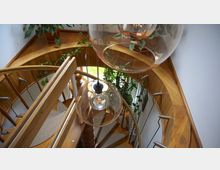 Holztreppe in spiralförmiger Bauweise mit Geländer aus Holz und Metall, umgeben von Topfpflanzen und einem hellen Interieur. Große, hängende Glühbirnen in Glaslampenschirmen sind im Vordergrund sichtbar.