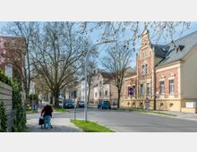 Straßenszene in einem Wohngebiet mit Reihenhäusern, einem historischen Gebäude mit Ornamentfassade rechts und mehreren Bäumen entlang des Gehwegs. Im Vordergrund geht eine ältere Person mit einem Rollator auf einem gepflasterten Bürgersteig.