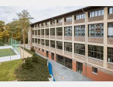 Außenansicht eines mehrstöckigen Gebäudes mit Backsteinfassade und großen Fenstern, flankiert von einem gepflasterten Weg und begrünten Bereichen. Links im Bild befindet sich ein eingezäuntes Sportfeld mit Kunstrasen, umgeben von Bäumen.