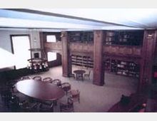 Ein Leseraum mit Holzmöbeln in einer Bibliothek. Zu sehen sind ein runder Tisch mit Stühlen, eine umlaufende Galerie mit Bücherregalen und große Fenster, die Tageslicht hereinlassen.