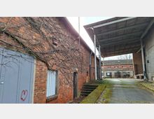 Schmaler Hof zwischen Backstein-Industriegebäuden mit überwucherten Ranken an der Wand, verschlossenen Türen und kleinen Fenstern. Rechts überspannt ein großes Vordach aus Metall und Holz die asphaltierte Zufahrt; im Hintergrund stehen weitere Lagergebäude.
