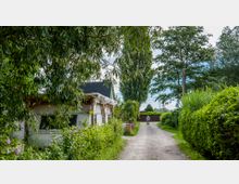 Ein ländlicher Weg mit Kiesbelag, gesäumt von dichtem grünen Gebüsch und Bäumen. Links steht ein weißes Gebäude mit dunklem Dach und Holzkonstruktion, im Hintergrund fährt eine Person mit einem Fahrrad in Richtung eines geschlossenen Holztors.