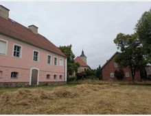 Ein ländlicher Hof mit einem rosa verputzten Gebäude links, einer Kirche mit Glockenturm im Zentrum und einem roten Backsteinhaus rechts. Im Vordergrund liegt geschnittenes Heu auf einer Wiese, umgeben von einzelnen Bäumen.