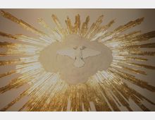 Relief einer weißen Taube, die von einer weißen Wolke umgeben ist, mit goldenen Strahlen, die kreisförmig vom Zentrum ausgehen. Das Kunstwerk symbolisiert himmlische oder spirituelle Themen.