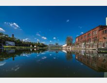 Backsteinbauten und ein modernes Glashaus stehen an einem ruhigen Gewässer, das die Umgebung klar spiegelt. Im Hintergrund sind Bäume, ein Industrie-Schornstein sowie eine blaue Himmelslandschaft mit kleinen Wolken zu sehen.