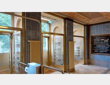 Eingangsbereich eines historischen Schwimmbads mit großen Glastüren, blauen Fliesenwänden und Heizkörpern. Rechts hängt eine Tafel mit Informationen zu Öffnungszeiten und Badeangeboten.