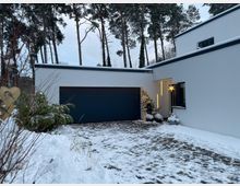 Moderner, weißer Flachdach-Bungalow mit großer dunkler Garagentür und gepflasterter, teilweise verschneiter Einfahrt. Im Hintergrund stehen hohe Kiefern, am Hauseingang ist eine beleuchtete Stern-Dekoration zu sehen.