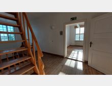 Wohnraum mit hölzernem Treppenaufgang im Vordergrund und hellem Zimmer im Hintergrund. Die Räume haben einen Holzboden, weiße Wände und große Fenster, durch die Tageslicht einfällt.