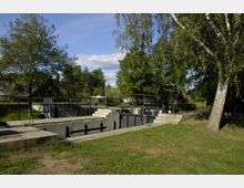 Schleusenanlage in einer grünen Parklandschaft mit Bäumen und Wiese. Betonwände und Metallgeländer umgeben die Schleuse, die von Spazierwegen eingefasst ist.