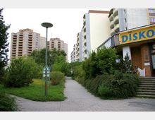 Eine Wohngegend mit mehrstöckigen Hochhäusern im Hintergrund, umgeben von dichter Vegetation. Im Vordergrund befinden sich ein gepflasterter Gehweg, ein Wegweiser und ein Gebäude mit einem gelben Schild mit der Aufschrift "Die Disko".