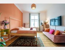 Helles Schlafzimmer mit einer orangefarbenen Akzentwand, einem Holzbett mit passendem Nachttisch, einer rosa Couch und einem gemusterten Teppich auf Holzdielenboden. Ein großes Fenster mit grünen Vorhängen und ein an der Wand montierter Fernseher ergänzen die Einrichtung.
