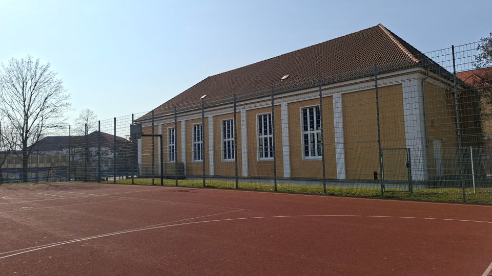 Eingezäunter Sportplatz mit rotem Belag vor einem langen, gelb-weißen Gebäude mit Satteldach und hohen, schmalen Fenstern. Im Hintergrund stehen weitere Gebäude und kahle Bäume unter klarem Himmel.