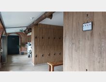 Ein Innenraum mit Holzschränken an der rechten Wand, einer Holzbank im Vordergrund und Backsteinwänden. Im Hintergrund befindet sich eine schwarze Tür mit der Aufschrift „Umkleiden“, daneben stehen Pflanzen und es hängt ein Schild an der Wand.