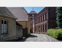 Gepflasterte kleine Gasse mit Gebäuden aus unterschiedlichen Materialien: links ein verblasstes Haus mit Ziegeldach, rechts ein rotes Backsteingebäude mit zahlreichen Fenstern. Im Hintergrund ist ein Kirchturm mit Uhr und spitzem Dach zu sehen.