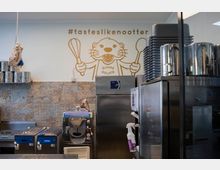 Innenraum einer Gelateria mit moderner Küchenausstattung, darunter Eismaschinen und Edelstahlgeräte. Eine Wand zeigt eine Illustration eines Otters mit Schneebesen und Löffel sowie den Schriftzug "#tasteslikenootter".