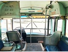 Innenansicht eines klassischen Busses mit grünlicher Farbgebung. Sicht auf den Fahrersitz, das Lenkrad, die Armaturen sowie die vordere Einstiegstür und Teil der Passagiersitze.