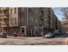 Straßenecke mit einem mehrstöckigen Wohngebäude aus Putzfassade und Balkonen. Im Erdgeschoss befindet sich ein Café mit Außenbestuhlung und beschrifteten Tafeln, daneben sind geparkte Autos und Fahrräder auf Kopfsteinpflasterstraßen sichtbar.