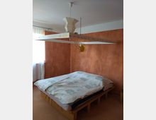 Schlafzimmer mit einem Holzbett und einer bunt gemusterten Bettdecke, umgeben von orangefarben gestrichenen Wänden. Eine große, ungewöhnliche Hängelampe mit dekorativen Elementen hängt zentral über dem Bett; links befindet sich ein teilweise verdecktes Fenster mit einem weißen Vorhang.