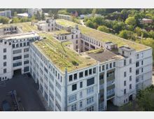 Luftaufnahme eines großen, mehrstöckigen Industriegebäudes mit heller Fassade und zahlreichen Fenstern. Das Flachdach ist begrünt, und die Umgebung zeigt Bäume, Wohngebäude und kleinere Industrieanlagen.