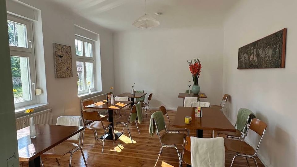 Heller Café- oder Bistro-Innenraum mit hohen weißen Wänden, großen Fenstern und poliertem Holzdielenboden. Mehrere kleine Tische mit Metall- und Holzstühlen stehen verteilt; an der Wand hängen Bilder, und auf einem Tisch steht eine Vase mit roten Blumen.