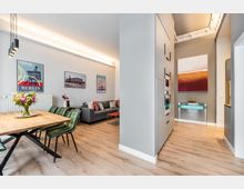 Offener Wohn‑ und Essbereich in einer modernen Wohnung mit Holzboden, Esstisch mit grünen Stühlen, grauem Sofa und gerahmten Wandbildern. Rechts führt ein Flur mit hohen Einbauschränken und integrierten Geräten zu einem Konsolentisch unter einem großen abstrakten Gemälde.