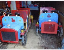Zwei bunt bemalte, altmodische Kinderfahrzeuge in rot und blau stehen nebeneinander in einer Garage oder einem Lagerraum. Im Hintergrund sind weitere Gegenstände und Fahrzeuge erkennbar.