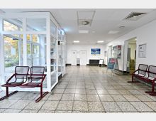 Heller Flur in einem öffentlichen Gebäude mit beigen Bodenfliesen, weißen Wänden und mehreren roten Metallstühlen. Auf der rechten Seite befinden sich Infotafeln und ein Zugang zu weiteren Räumen, links eine verglaste Eingangstür mit Blick nach draußen.