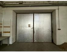 Innenansicht eines industriellen Lager- oder Garagenraums mit großer zweiflügeliger Metalltür in einer Wandnische. Sichtbare Deckenrohre und Kabelkanäle sowie schlichte weiße Wände und Betonboden prägen den Raum.