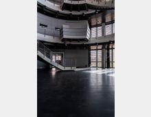 Großzügiger, moderner Innenraum mit dunklem Steinboden, geschwungenen Metallelementen und einer breiten Treppe mit Geländer aus Metall. Die Wände und Fensterfronten lassen Tageslicht einströmen, und es sind mehrere Balkone und Geländer auf verschiedenen Etagen sichtbar.