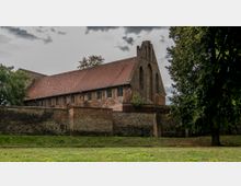 Backstein-Gebäude mit spitzem Giebel, umgeben von einer historischen Backstein- und Feldsteinmauer, im Vordergrund eine Grünfläche mit Bäumen. Der Himmel zeigt graue Wolken.