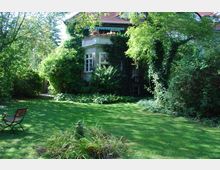 Ein Haus mit begrüntem Balkon, umgeben von dichtem Bewuchs und einem gepflegten Garten mit Rasenfläche. Im Vordergrund eine kleine Statue und eine Holzbank, im Hintergrund viele Bäume und Sträucher.