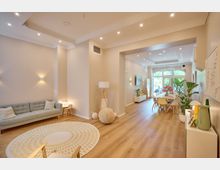 Helles, modern eingerichtetes Wohnzimmer mit einem offenen Essbereich. Holzboden, beige Wände, skandinavisches Mobiliar, eine große Fensterfront im Hintergrund sowie dekorative Pflanzen und ein runder Teppich prägen den Raum.