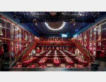 Innenraum eines Theaters mit festlichem Ambiente. Der Raum verfügt über rote Wände, Galerien auf mehreren Ebenen, runde Tische mit roten Tischdecken, eine geschwungene Treppe und eine beleuchtete Deckeninstallation. Poster und Kostüme schmücken die Wände.