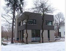 Modernes zweistöckiges Wohnhaus mit flachem Dach und einer Fassade in Beige und Dunkelgrau, umgeben von schneebedecktem Boden und kahlen Bäumen. Im Vordergrund verläuft ein rot-weißes Absperrband.