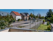 Straßenszene in einem Wohngebiet mit Einfamilienhäusern, einem Gehweg und einer Bushaltestelle mit einem überdachten Wartehäuschen. Vor den Häusern sind Rasenflächen und Zäune sichtbar, die Straße ist von Bäumen gesäumt.