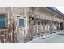 Holzgebäude mit verwitterten, weißen Türen und Fenstern, die kleine Glasscheiben enthalten. Die Fassade zeigt deutliche Altersspuren, mit abgeblätterter Farbe und freiliegendem Holz, auf einem überdachten Hofbereich mit Stützpfeilern.