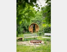 Holzhütte in Form eines Fasses inmitten eines grünen Gartens, umgeben von Bäumen und Sträuchern. Im Vordergrund stehen Bänke um eine Feuerstelle, die von Steinen eingefasst ist.