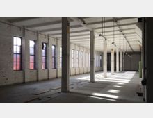 Großer, leerer Innenraum eines ehemaligen Industriegebäudes mit weiß gestrichenen Ziegelwänden, großen vertikalen Fenstern und einer Decke mit sichtbaren Trägern. Der Boden ist unvollständig, und mehrere Säulen verlaufen in einer geraden Linie durch den Raum.