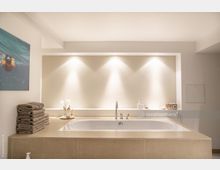 Modernes Badezimmer mit einer eingelassenen Badewanne aus beigem Stein, flankiert von Deko-Elementen wie Handtüchern, Flaschen und Accessoires. Die Wand dahinter ist in warmem Licht beleuchtet und minimalistisch gestaltet, mit einem Bild an der linken Wand.