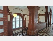 Innenansicht eines historischen Flurs mit roten Backsteinpfeilern und Rundbögen, einer großen bogenförmigen Fensteröffnung und hängenden Kugelleuchten. Ein gemusterter Fliesenboden und ein schmiedeeisernes Geländer säumen die Galerie entlang der Wand.