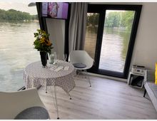 Wohnraum mit Blick auf einen See durch große Fenster. Ein runder Tisch mit gemusterter Tischdecke und Blumenvase steht zwischen zwei weißen Stühlen; im Hintergrund sind ein Sofa, ein kleiner Beistelltisch und ein wandmontierter Fernseher erkennbar.