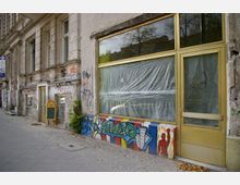Fassade eines alten Gebäudes in urbaner Umgebung mit großen, vergilbten Fensterscheiben, die teils mit Folie abgedeckt sind. Teile der Fassade sind mit bunten Graffiti und Verwitterungsspuren versehen, während der Gehweg aus Kopfsteinpflaster besteht.